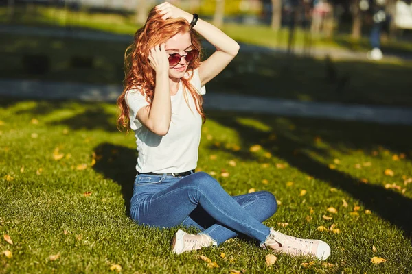 Een mooie roodharige meisje, opgewarmd door de stralen van de zon, zit op een gazon en poseren voor de camera. Het meisje draagt een T-shirt met jeans, een bril op haar gezicht en een moderne gadget op haar arm — Stockfoto