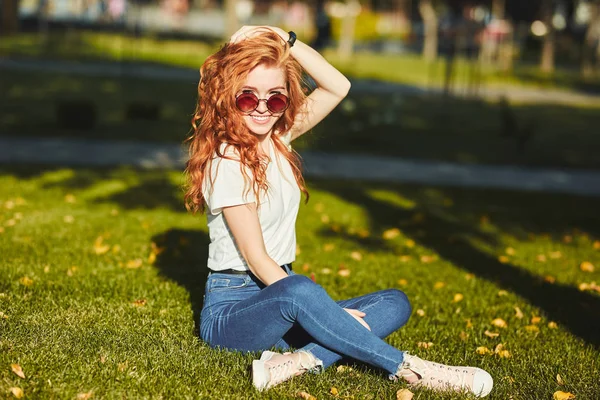 Une jolie fille aux cheveux roux, réchauffée par les rayons du soleil, est assise sur une pelouse et pose pour la caméra. La fille porte un T-shirt avec un jean, des lunettes sur son visage et un gadget moderne sur son bras — Photo