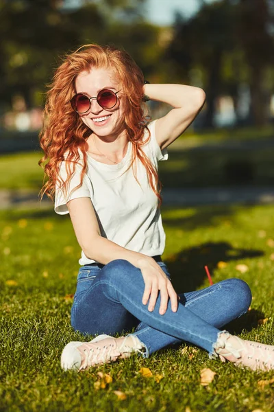 Una encantadora chica pelirroja, calentada por los rayos del sol, está sentada en un césped y posando para la cámara. La chica lleva una camiseta con jeans, gafas en la cara y un gadget moderno en el brazo. —  Fotos de Stock