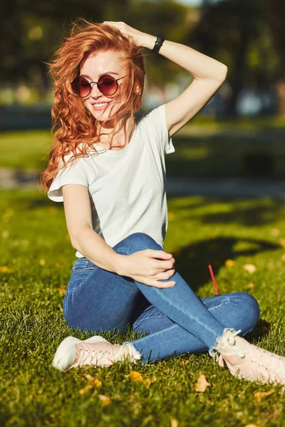 Una encantadora chica pelirroja, calentada por los rayos del sol, está sentada en un césped y posando para la cámara. La chica lleva una camiseta con jeans, gafas en la cara y un gadget moderno en el brazo. —  Fotos de Stock