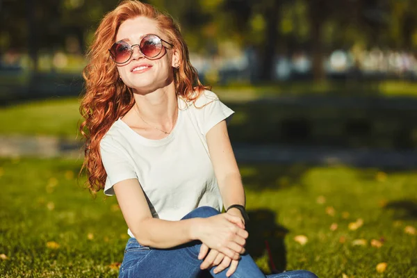 Güzel bir kızıl saçlı kız, güneş ışınları tarafından ısınmış, bir çim üzerinde oturuyor ve kamera için poz. Kız kot pantolon, yüzünde gözlük ve kolunda modern bir alet olan bir tişört giyiyor. — Stok fotoğraf