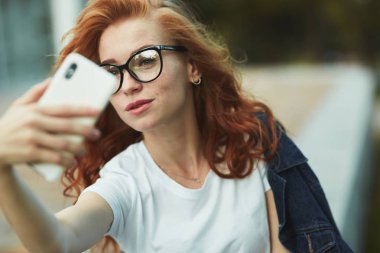 Güzel bir kızıl saçlı kız halka açık bir bahçede dinleniyor ve cep telefonuyla selfie çekiyor. Kız kot pantolon, yüzünde gözlük ve kolunda modern bir alet olan bir tişört giyiyor.