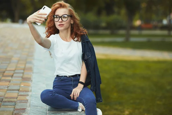 Piękna ruda dziewczyna odpoczywa w publicznym ogrodzie i zabiera selfie na jej telefon komórkowy. Dziewczynka ma na sobie koszulkę z dżinsami, okulary na twarzy i nowoczesny gadżet na ramieniu — Zdjęcie stockowe