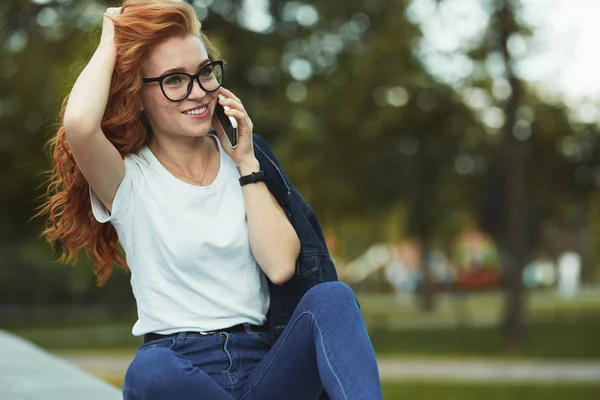 Vacker rödhårig flicka med en vacker figur och en telefon i hennes händer. Flickan kommunicerar på sin moderna Gadget med sina släktingar och vänner. Hon bär en T-shirt och jeans — Stockfoto