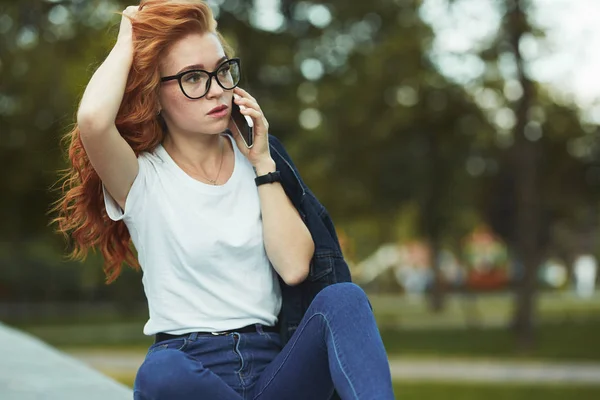 Vacker rödhårig flicka med en vacker figur och en telefon i hennes händer. Flickan kommunicerar på sin moderna Gadget med sina släktingar och vänner. Hon bär en T-shirt och jeans — Stockfoto