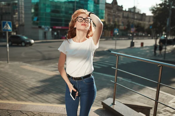 Mooie roodharige meisje staat in de straat op de achtergrond van de stad en poseren op camera. Het meisje draagt een T-shirt met jeans — Stockfoto