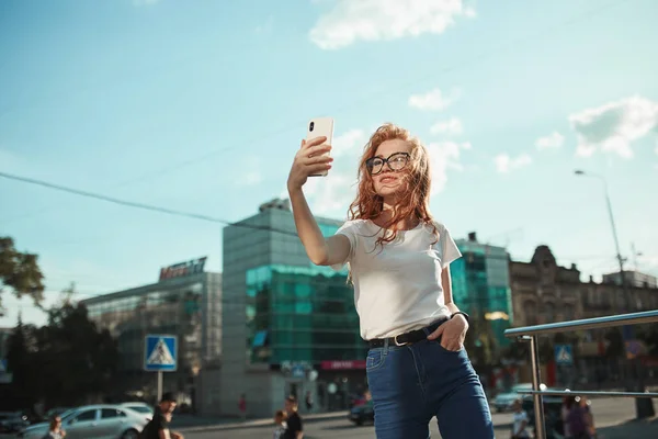 Красива руда дівчина стоїть на вулиці на фоні міста і бере селфі на свій мобільний телефон. Дівчина в футболці з джинсами — стокове фото