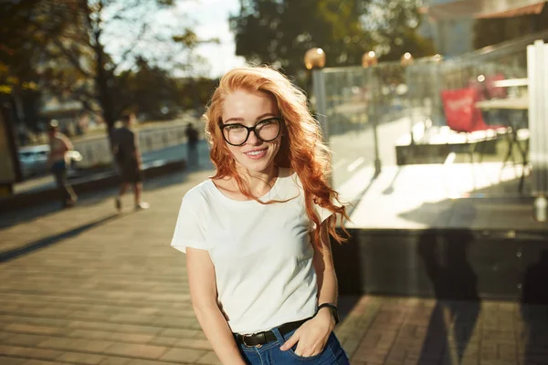 Vackra rödhårig flicka står på gatan på bakgrunden av staden och poserar på kameran. Flickan bär en T-shirt med jeans — Stockfoto