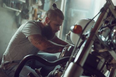 Yakışıklı sakallı adam garajda motosikletini tamir ediyor. Kot pantolon ve tişört giyen bir adam.