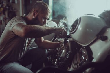 Yakışıklı sakallı adam garajda motosikletini tamir ediyor. Kot pantolon ve tişört giyen bir adam.
