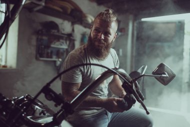 Yakışıklı sakallı adam bir motosiklet koltuğunda oturur ve uzağa bakar