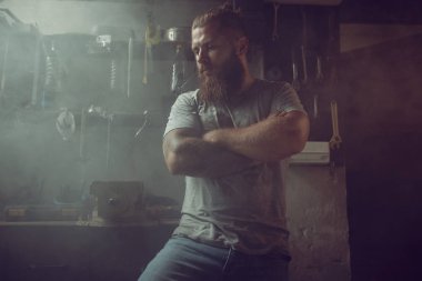 Tamir araçlarının arka planına karşı garajında duran sakallı yakışıklı vahşi adam