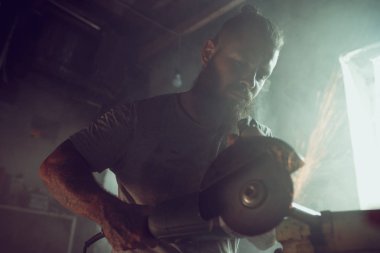 Yakışıklı vahşi erkek sakallı, garajında bir motosikleti tamir eden dairesel testereyle çalışıyor. Garajda bir sürü kıvılcım ve testere dumanı