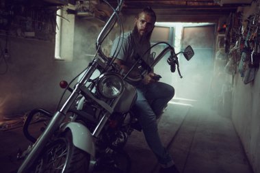 Handsome brutal man with a beard sitting on a motorcycle in his garage, wiping his hands and looking to the side clipart