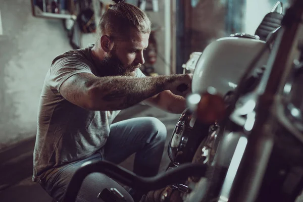 Bonito barbudo a reparar a mota na garagem. Um homem usando jeans e uma camiseta — Fotografia de Stock