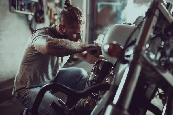 Un bel homme barbu réparant sa moto dans le garage. Un homme portant un jean et un t-shirt — Photo