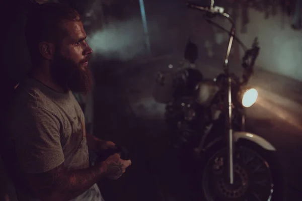 Bonito homem brutal com uma barba está de pé em sua garagem contra o fundo de uma motocicleta e olhando para longe — Fotografia de Stock