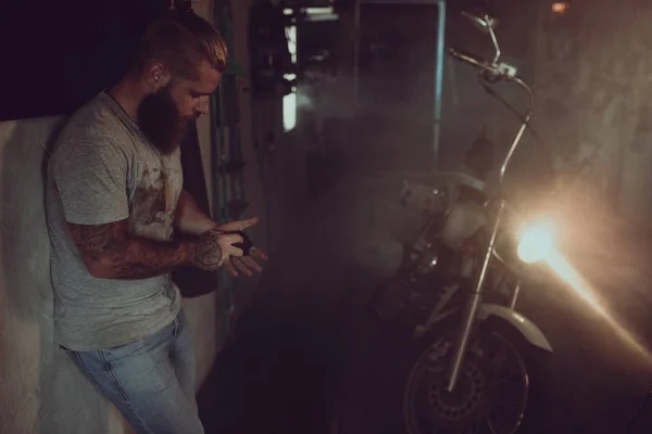 Bonito homem brutal com uma barba está de pé em sua garagem contra o fundo de uma motocicleta e olhando para longe — Fotografia de Stock