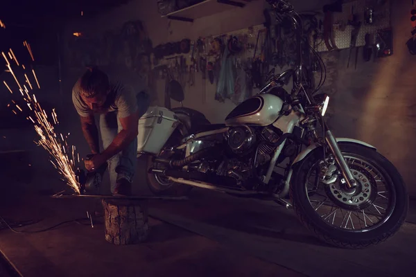 Bonito macho brutal com barba reparando uma moto em sua garagem trabalhando com uma serra circular. Na garagem um monte de faíscas e fumaça de serrar — Fotografia de Stock