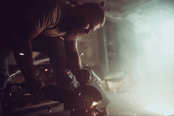 Bonito macho brutal com barba reparando uma moto em sua garagem trabalhando com uma serra circular. Na garagem um monte de faíscas e fumaça de serrar — Fotografia de Stock