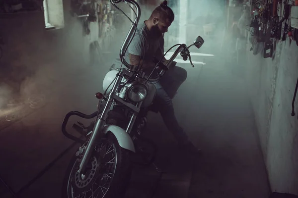 Όμορφος βίαιος άντρας με μούσι να κάθεται σε μια μοτοσικλέτα στο γκαράζ του, να σκουπίζει τα χέρια του και να κοιτάζει προς την πλευρά — Φωτογραφία Αρχείου