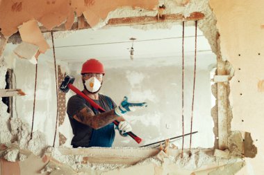 Elinde çekiçle inşaatçı beton duvarı yıkar inşaatçı koruyucu bir kostüm ve kask giyer.