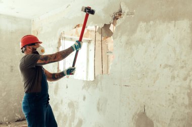 Elinde çekiçle inşaatçı beton duvarı yıkar inşaatçı koruyucu bir kostüm ve kask giyer.