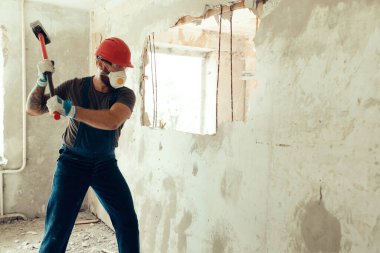 Elinde çekiçle inşaatçı beton duvarı yıkar inşaatçı koruyucu bir kostüm ve kask giyer.