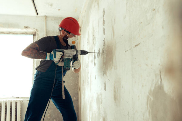 builder with perforator drills holes in concrete wall The builder is dressed in a protective suit and helmet