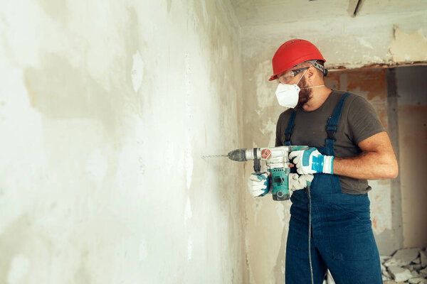 builder with perforator drills holes in concrete wall The builder is dressed in a protective suit and helmet