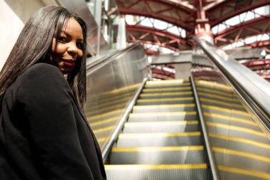 Uzun siyah saçlı ve siyah ceketli bir iş kadını modern bir ulaşım merkezinde yürüyen merdivene biniyor ve kendine güvenen bir ifadeyle geri dönüyor.