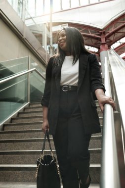 Modern bir binada el çantalı bir iş kadını merdivenlerden çıkıyor, korkulukları tutuyor ve neşeli bir gülümsemeyle uzaklaşıyor, kendine güveni ve zarafeti yayıyor