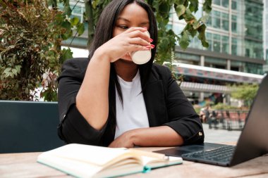 İş kadını dışarıdaki ahşap bir masada oturuyor. Yanında bir defterle dizüstü bilgisayarıyla kağıt bardaktan kahve içiyor.