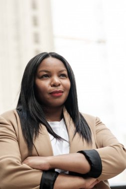 Başarılı bir iş kadını modern bir ofis binasında duruyor, kollarını kavuşturmuş özgüvenini sergiliyor ve düşünceli bir gülümseme, şirket dünyasında liderlik ve profesyonellik gösterisi yapıyor.