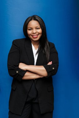 Siyah takım elbiseli bir iş kadını, kollarını mavi bir zemine doğru kıvırarak güvenle gülümsüyor, profesyonellik ve başarı yayıyor.