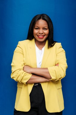 Başarılı bir iş kadını, canlı bir mavi arkaplan karşısında dururken kendine güveni tam, kollarını kavuşturmuş ve yüzünü aydınlatan sıcak bir gülümseme...