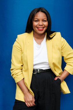 Sarı ceket ve siyah pantolon giyen kendine güvenen iş kadını, bir eli cebinde canlı mavi bir arka fona karşı poz verirken gülümsüyor.