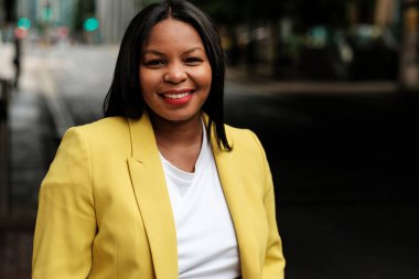 Kendine güvenen iş kadını, bulanık bir şehir ortamında canlı sarı bir ceket ve beyaz bir tişört giyerek profesyonelliği ve başarıyı yansıtan bir şekilde gülümsüyor.