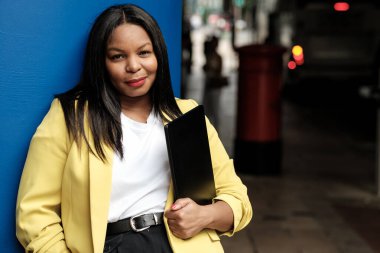 Sarı ceketli bir iş kadını, canlı mavi bir duvarın yanında elinde dizüstü bilgisayarla duruyor. Kendine güveni ve profesyonelliği var.