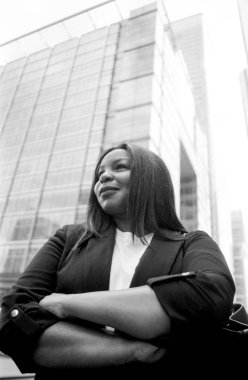 Modern bir ofis binasının önünde kollarını kavuşturmuş, özgüven ve profesyonellik saçan bir iş kadını tasvir edilmiş siyah beyaz analog bir görüntü.