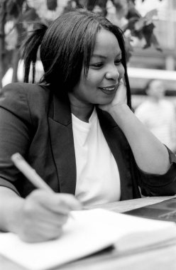 Bir iş kadınının açık bir kafedeki masada otururken defterdeki kalemle gülümseyen ve notlar alan tek renkli kareli analog görüntüsü.