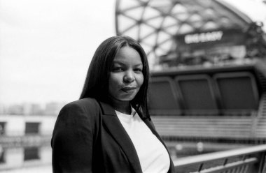 Modern bir şehir ortamında duran bir iş kadınının siyah ve beyaz analog fotoğrafı, kendine güvenen ve stil sahibi bir varlık sergiliyor.