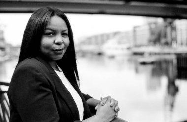 Tokalı elleri olan bir iş kadınının taneli tek renkli portresi. Arka planda bulanık bir kanal ve binalar olan şehir ortamında güvenle poz veriyor.