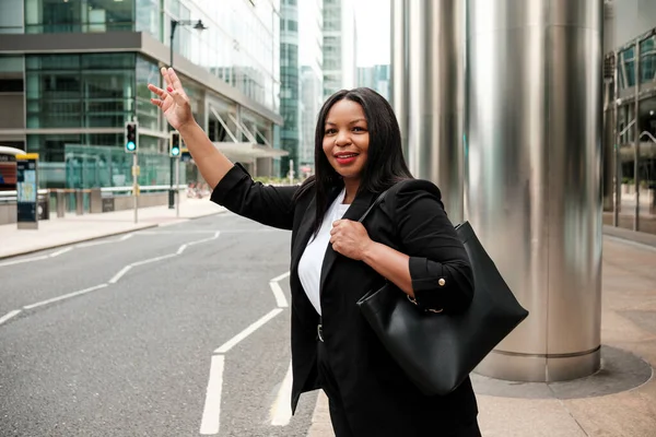 İş kadını, şehir caddesinde taksi çağırırken gülümser, siyah bir çanta taşır ve beyaz bir gömleğin üzerine siyah bir ceket giyer, arka planda modern ofis binaları vardır.