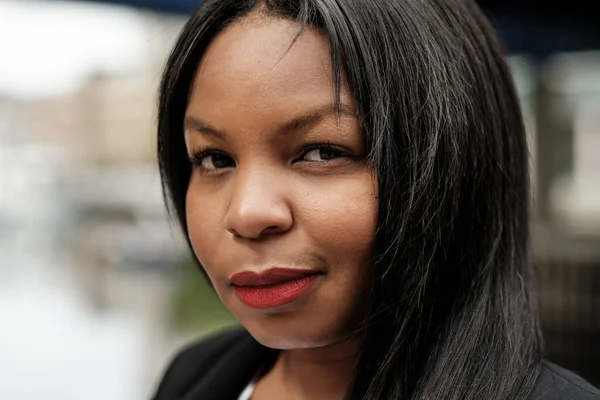 Uzun siyah saçlı ve çarpıcı kırmızı rujlu Afro-Amerikalı genç bir iş kadını, kendine güvenen bir ifadeyle dışarıda poz veriyor, canlı bir şehir ortamında profesyonellik ve hırs yayıyor.