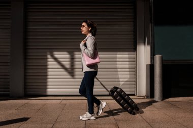 Woman traveler pulling luggage on wheels along an urban street pavement, moving towards adventure and new experiences under the contrasting shadows of a city building