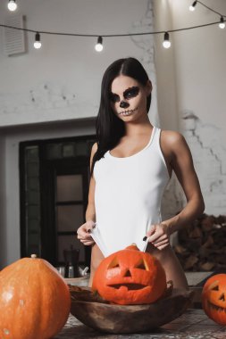 Genç ve güzel kadın, ürpertici makyaj giyiyor mayo ve uzun çorap ve kamera mutfak dolapları yakın dururken bakarak Cadılar Bayramı için dekore edilmiştir.