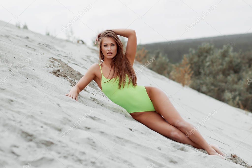 Seductive woman in stylish swimsuit kneeling in sand