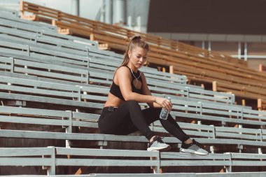 Kulaklıklı yorgun kadın tribünde dinleniyor ve su içiyor