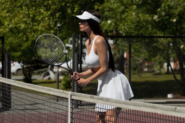 在当代体育俱乐部 穿着白色运动服和面容的性感年轻女运动员拿着球拍 靠在网球场上 带着诱惑力看着摄像机的侧视图 — 图库照片